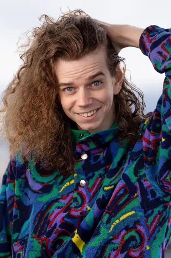 Smiling person with long curly hair and blue eyes, wearing colorful patterned fleece.