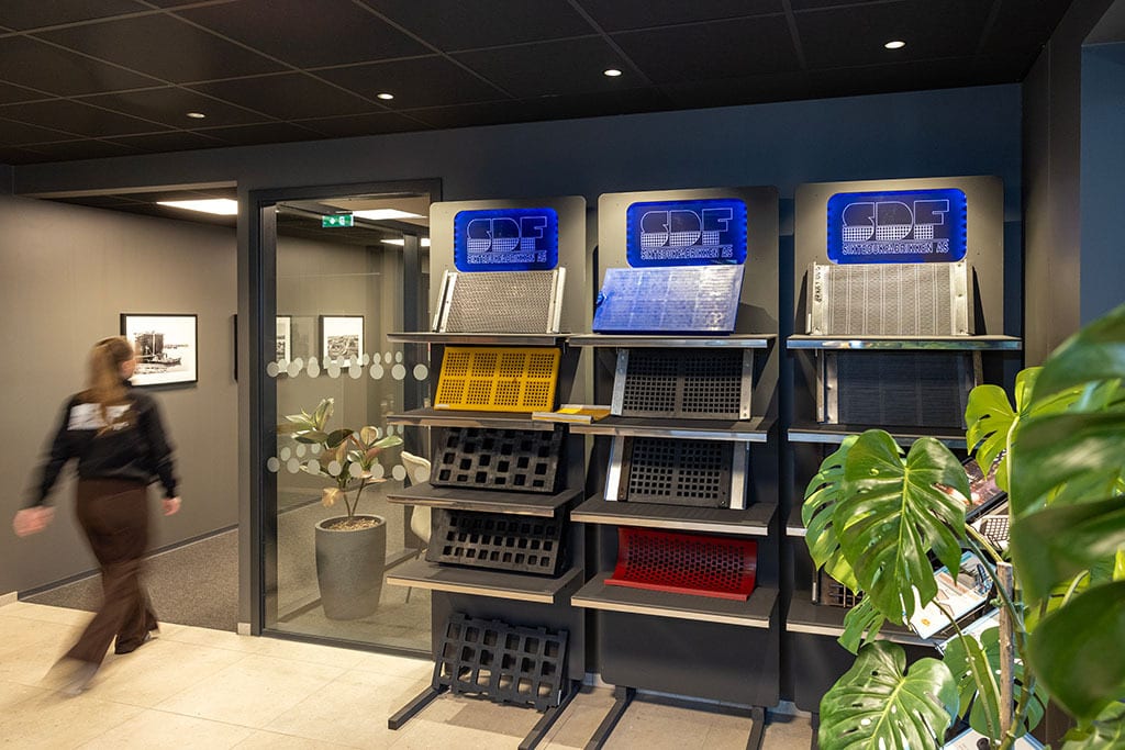 Showroom with colorful perforated panels on display, SDF logos, a blurred person, and lush plants.
