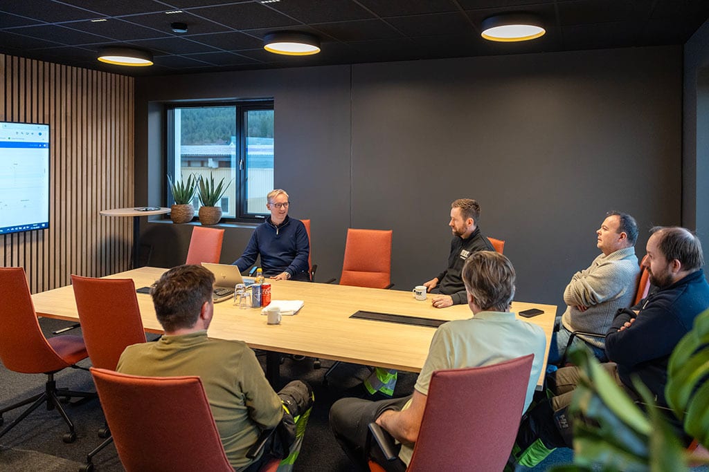 Six men in a modern meeting room, seated around a table with a laptop and screen.