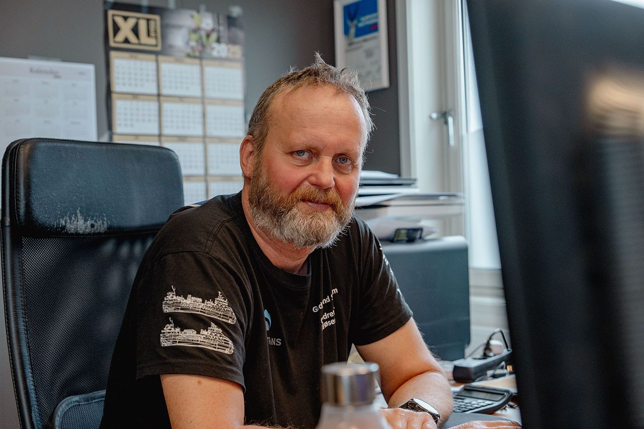 Man with beard in black t-shirt at office desk looks at camera.