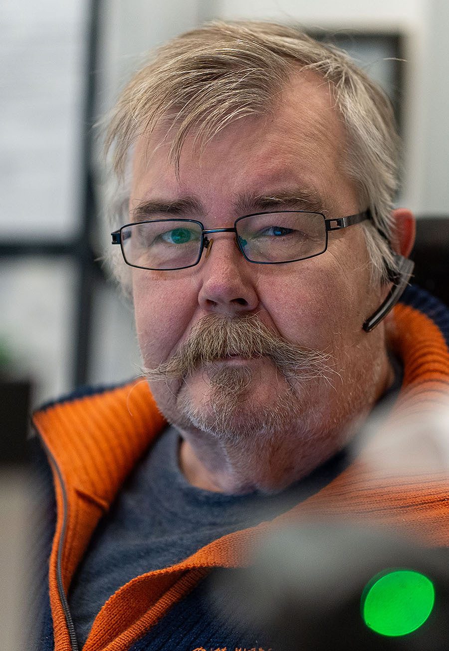 Older man with glasses, mustache, headset, and an orange and blue jacket.