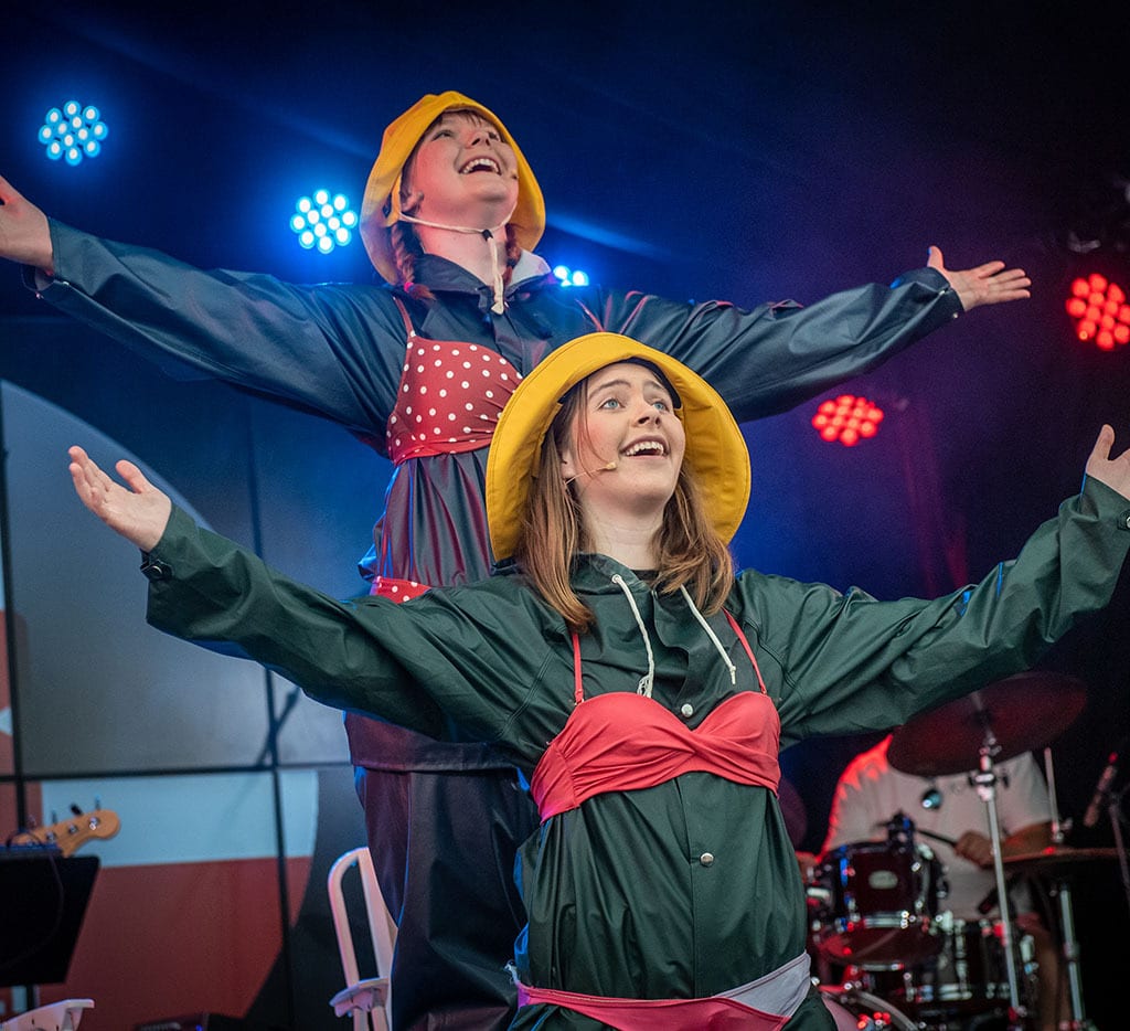 Two women on stage in raincoats and hats, smiling with arms outstretched under stage lights.