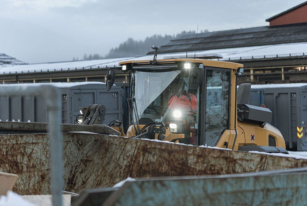 Yellow loader with bright lights, operator, and rusty bin in snowy industrial setting.