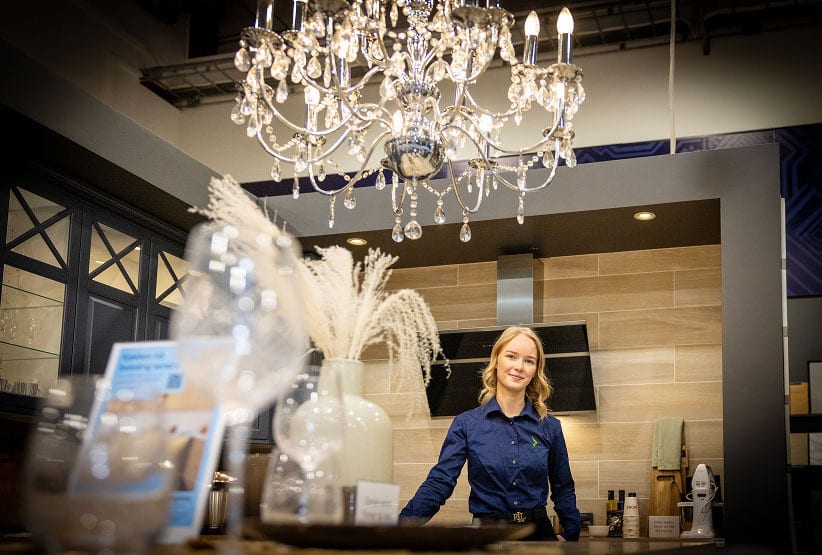 A smiling woman in a blue shirt stands in a modern kitchen showroom with a large crystal chandelier overhead.