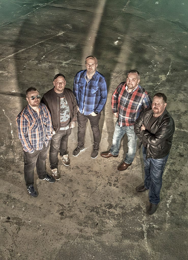 Five casually dressed men stand on a concrete floor looking up at the camera.