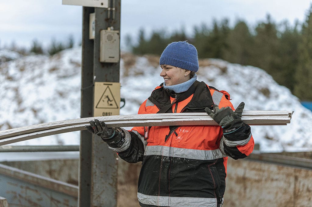 A smiling woman in winter work clothes carries metal bars outdoors in a snowy landscape.