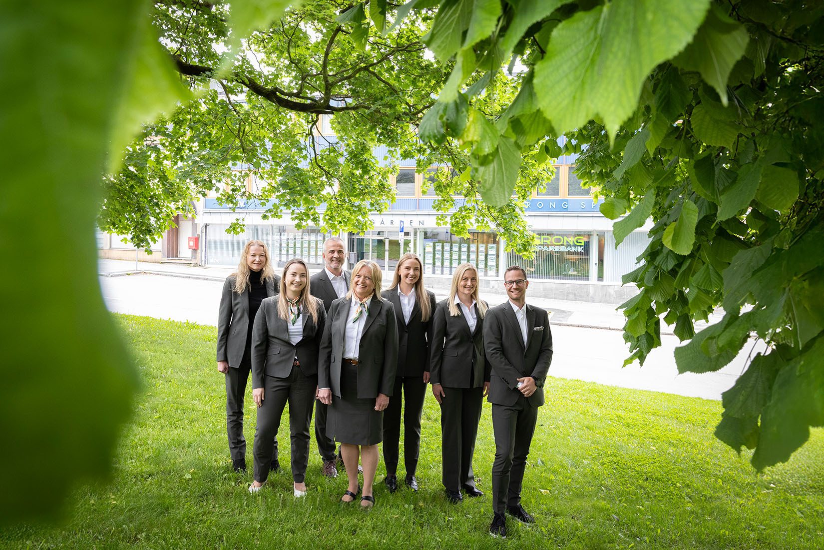 Seven people in business attire on grass, bank building and green leaves in view.