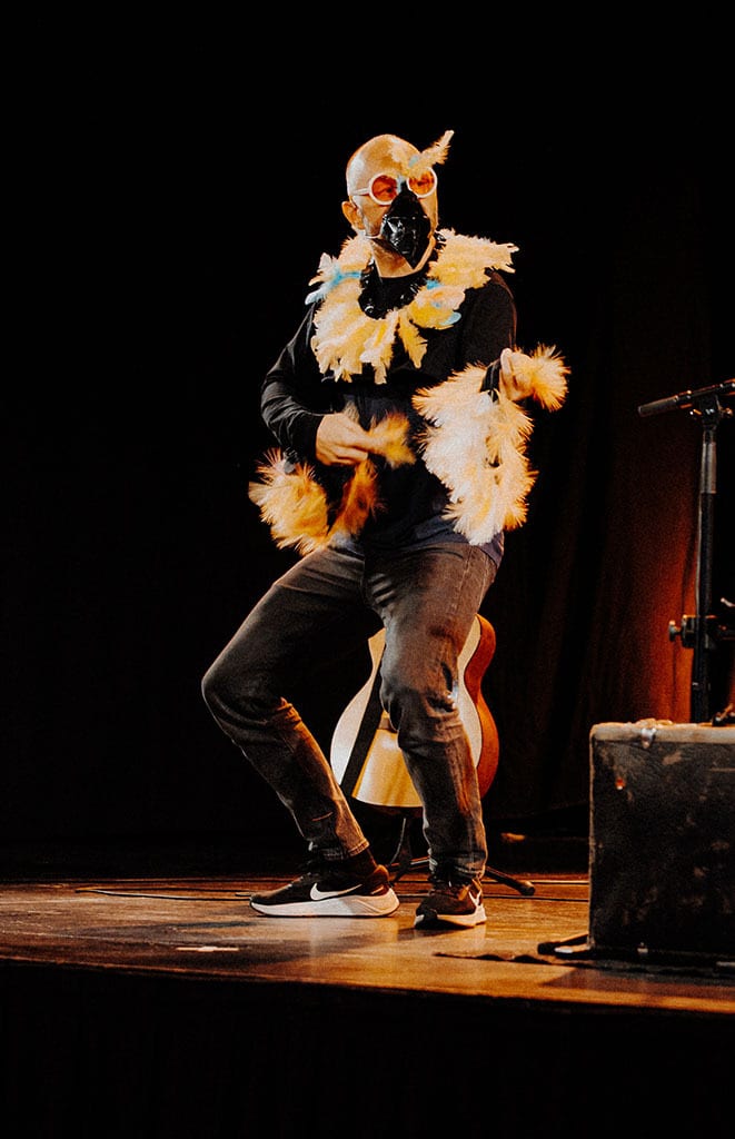 A person in a bird mask, orange glasses, and feather boa performs on stage, a guitar nearby.