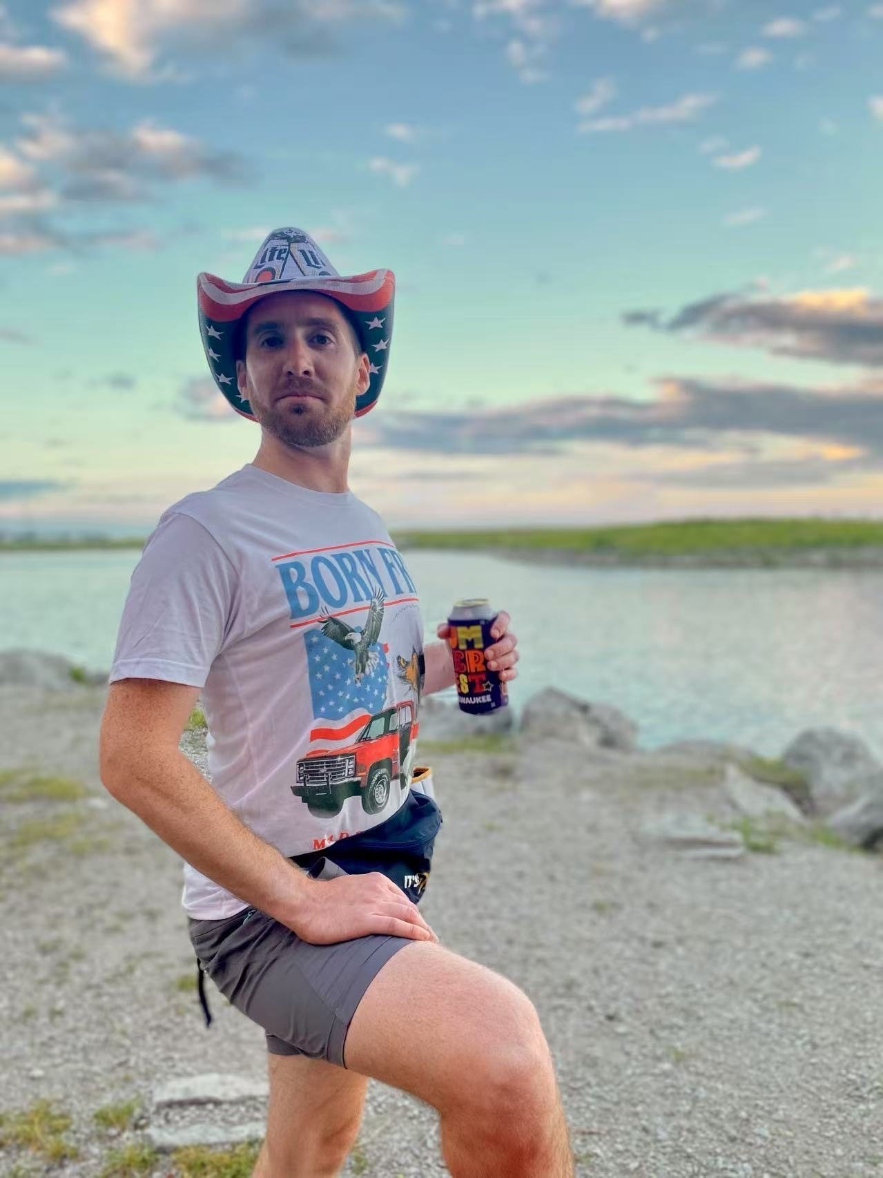 Man in Miller Lite cowboy hat and American flag shirt, holding a can by water.
