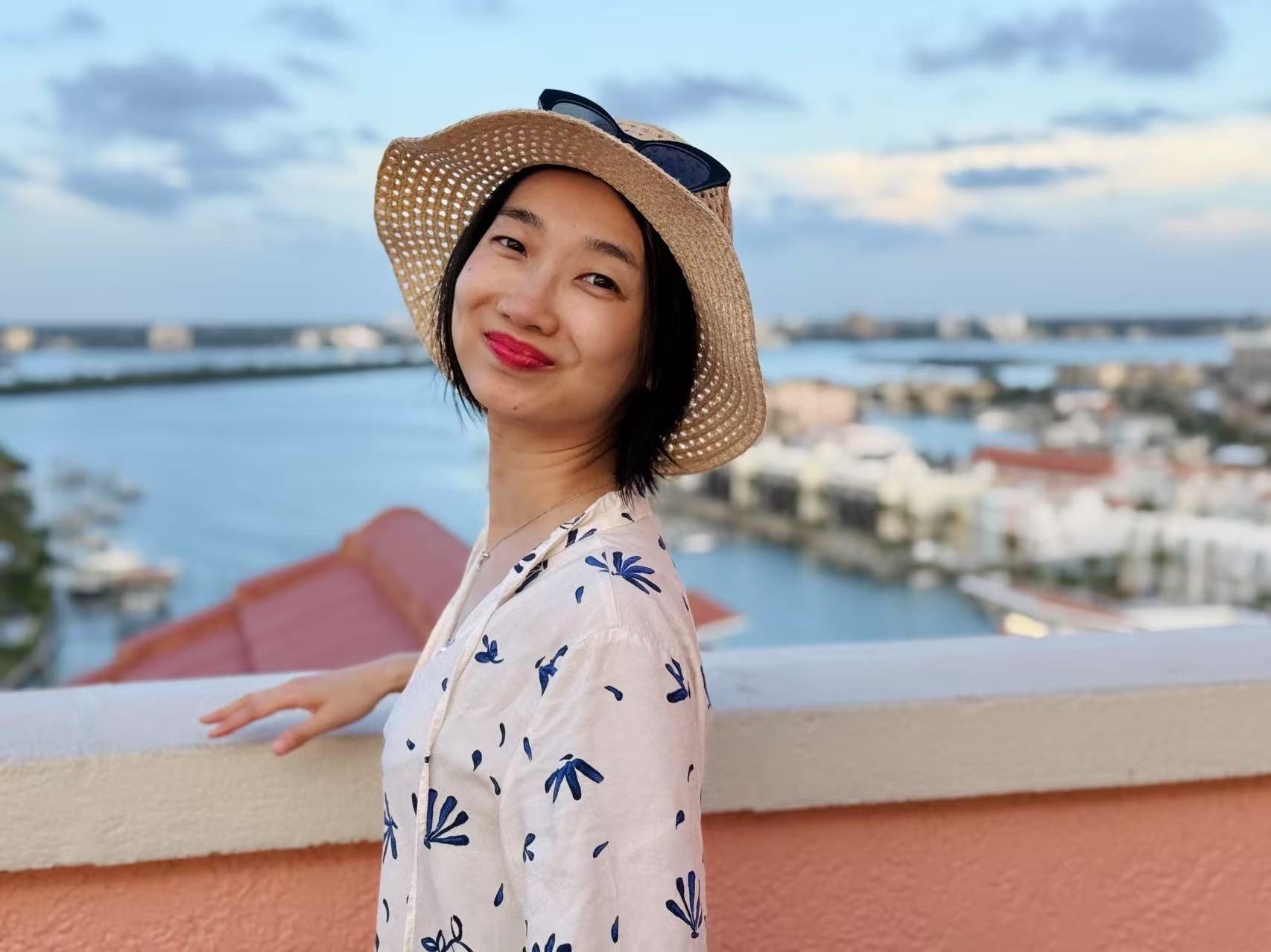 Smiling person in a straw hat on a balcony overlooking a waterfront city.