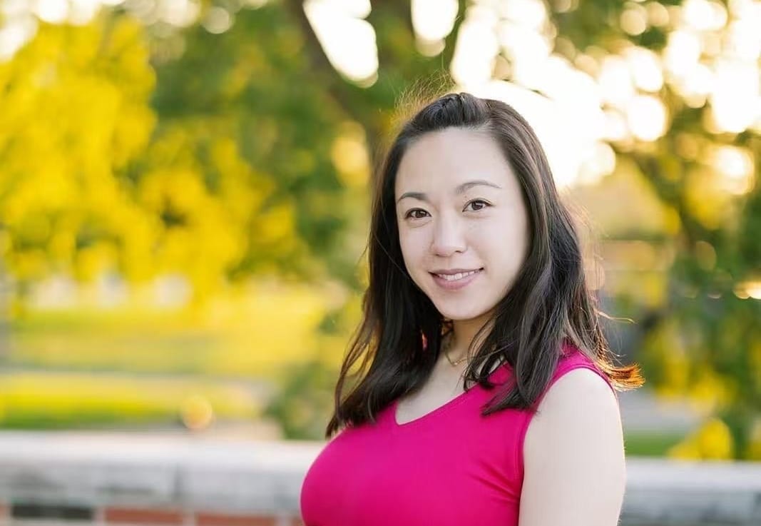 Smiling woman in pink top, outdoors with blurred yellow foliage.