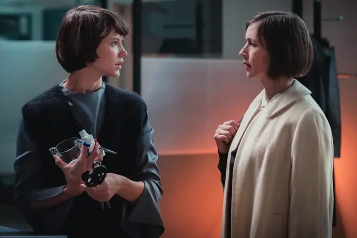 Two women with bob haircuts face each other, one holding a glass and tools, the other in a cream coat.