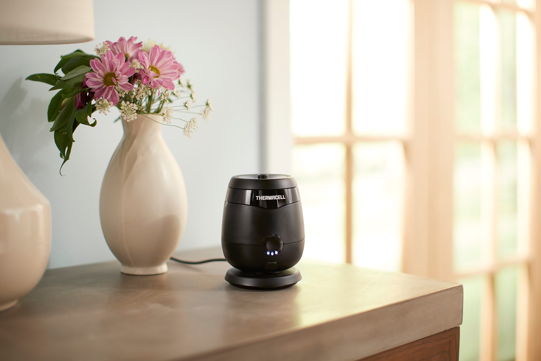 Black Thermacell mosquito repeller on a table next to a vase of pink flowers by a window.