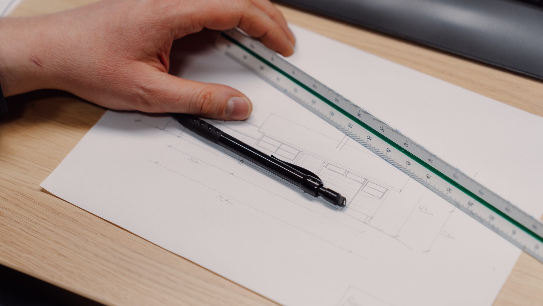 Hand holding a ruler on a drawing with a pen, showing architectural plans with measurements.