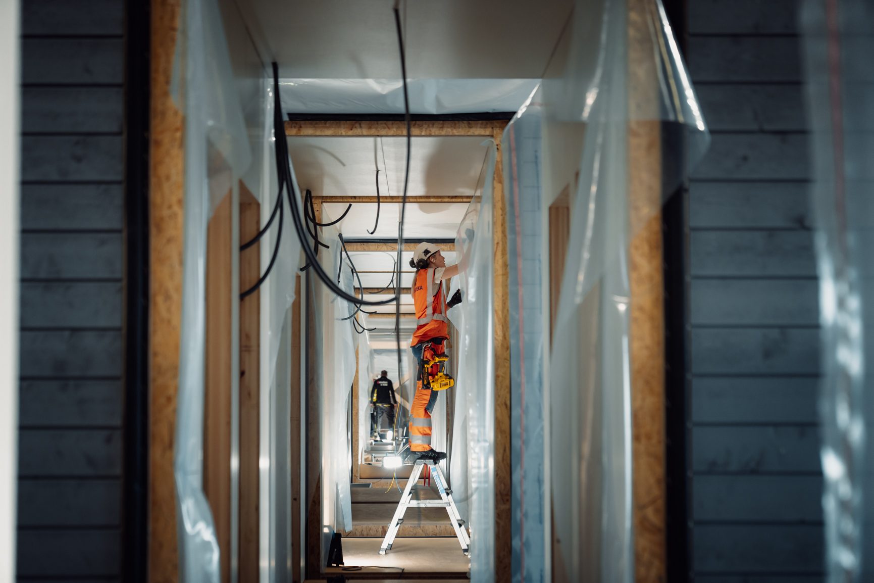 A construction worker on a ladder in a framed building hallway, installing overhead.
