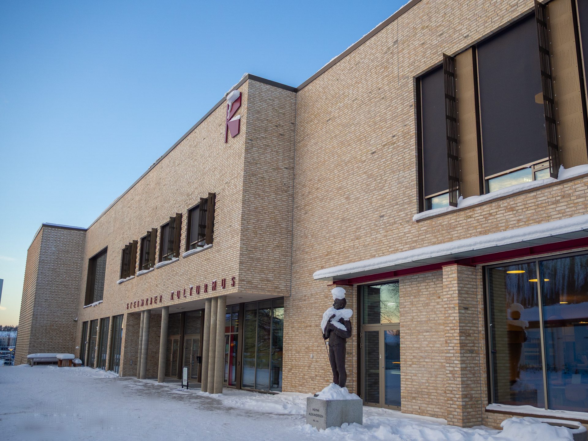 A snow-covered brick building named STEINKJER KULTURHUS with a statue in front under a clear blue sky.
