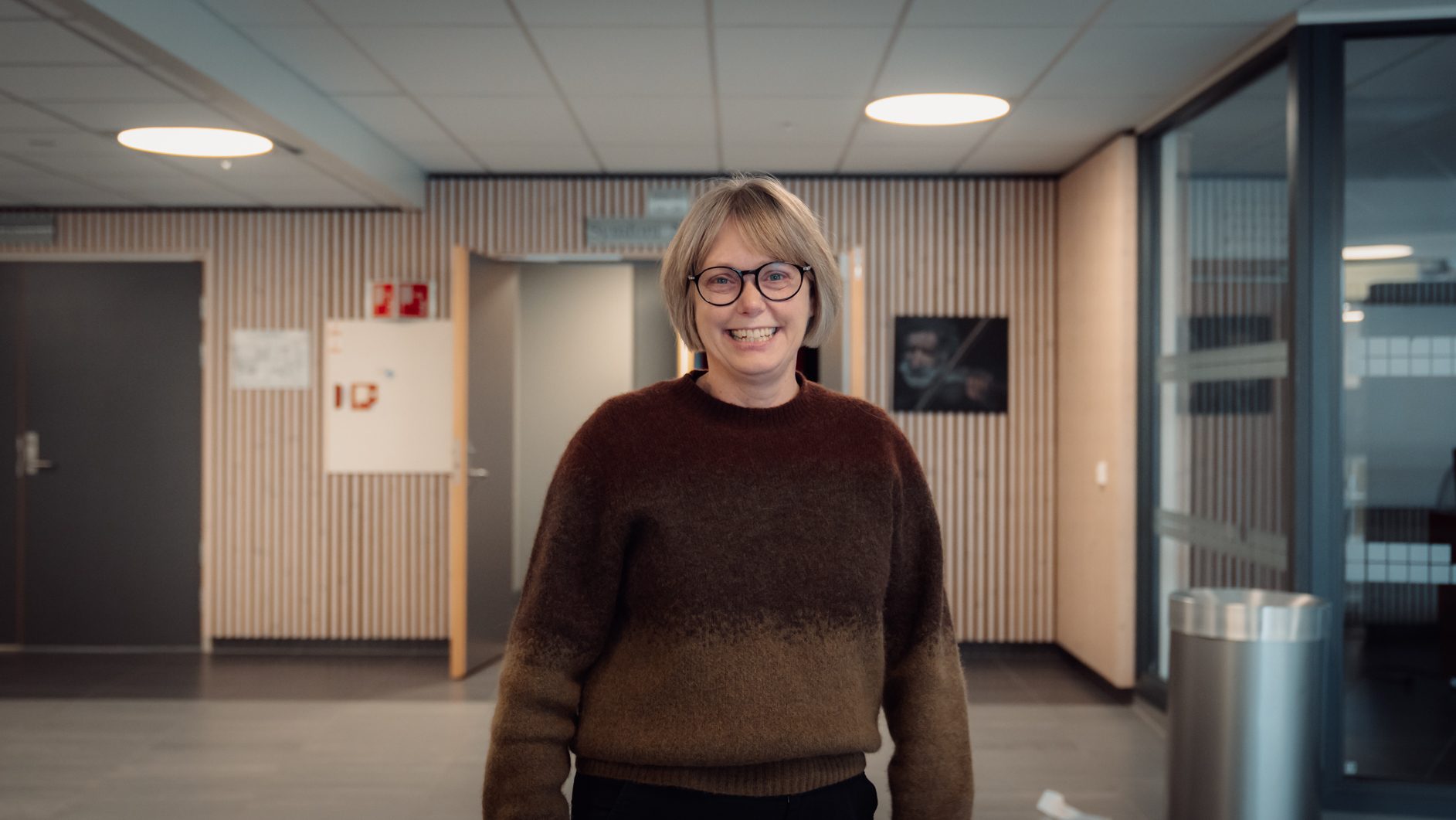 A smiling woman with short hair and glasses wearing a brown ombré sweater stands in a modern hallway.