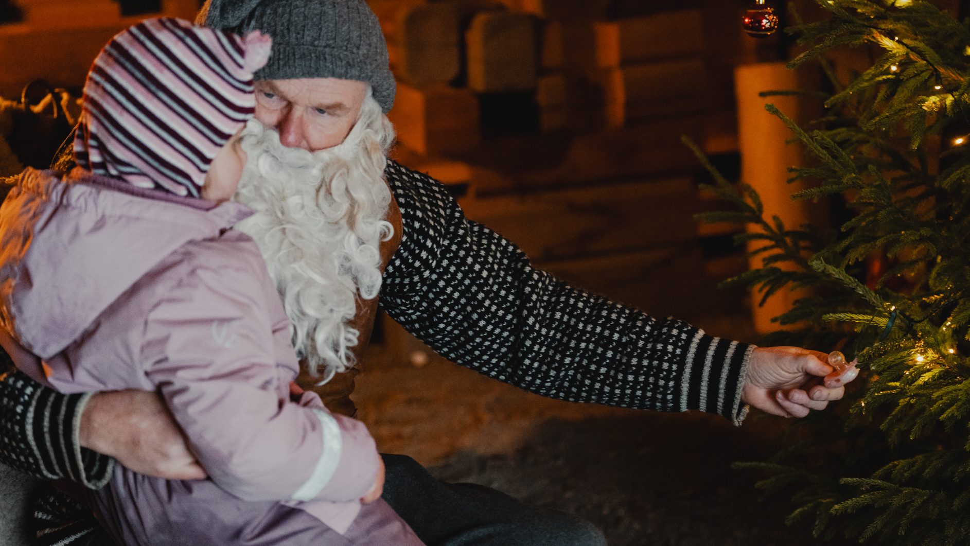 Santa holds a child, pointing at lights on a Christmas tree.
