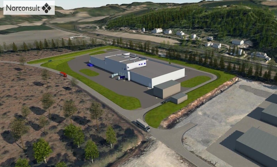 Aerial view of a white industrial building surrounded by green, with roads, trucks, and distant hills/houses.