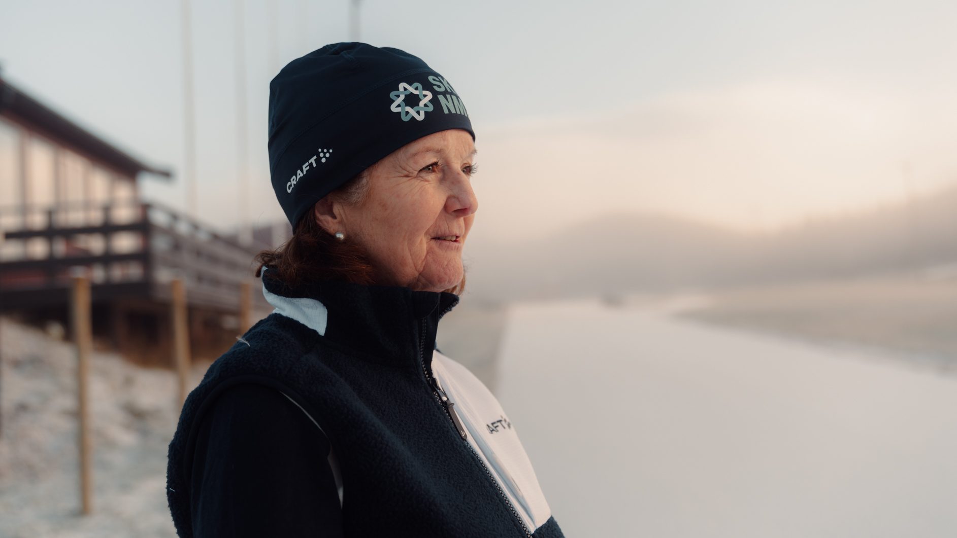 A smiling woman in a beanie and fleece jacket looks out over a misty, snowy winter scene.
