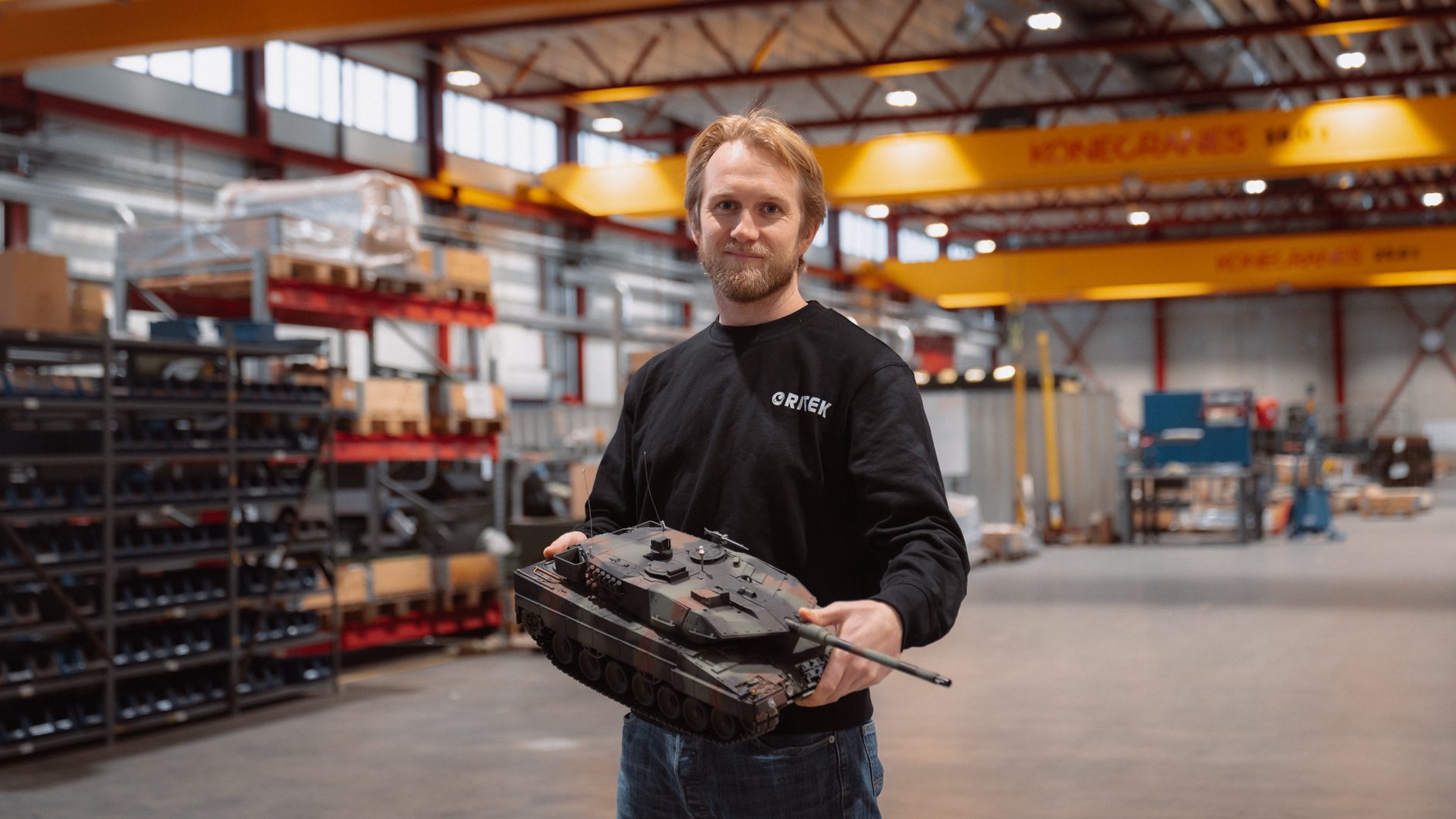 Man holding a camouflage model tank in a factory setting with shelves and overhead cranes.