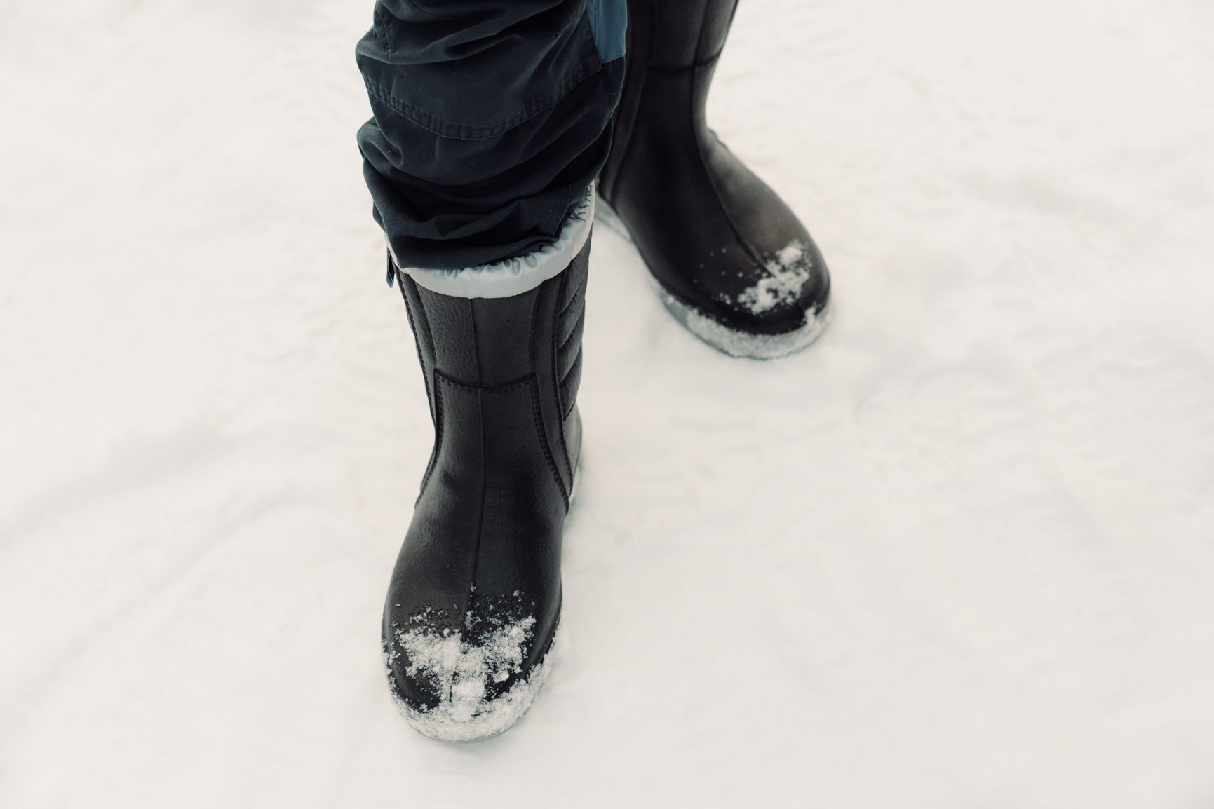 A low-angle shot of a person's legs wearing dark snow boots and pants, standing in fresh white snow.