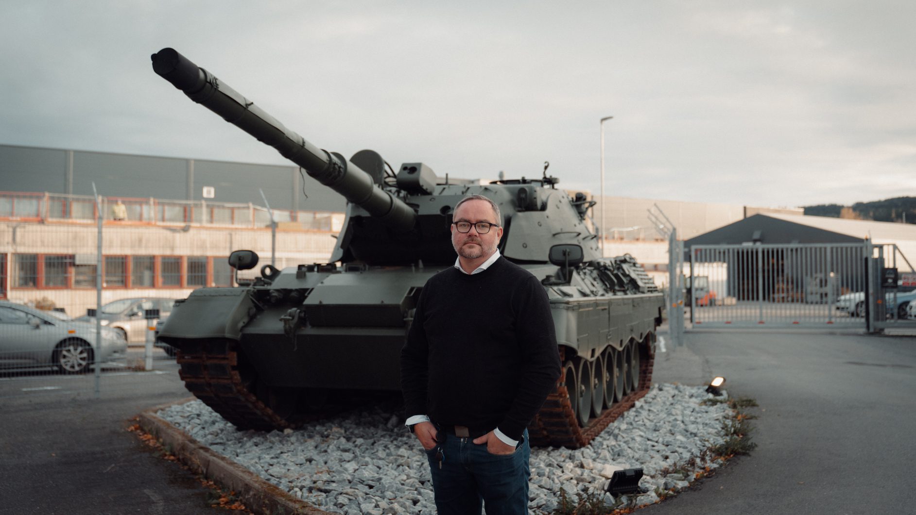 A man stands in front of a military tank at an industrial site.