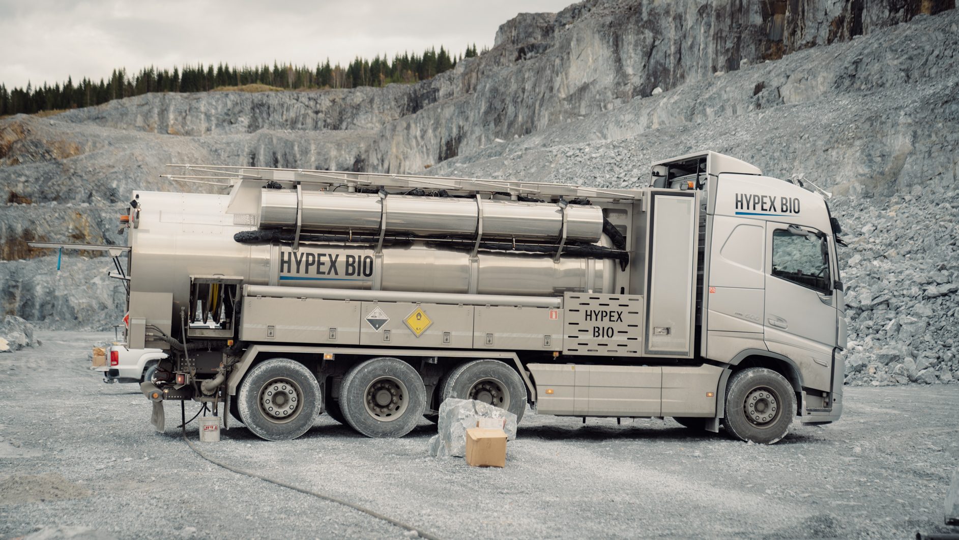 Silver HYPEX BIO tanker truck parked in a rocky quarry, with cliffs and pine trees in background.