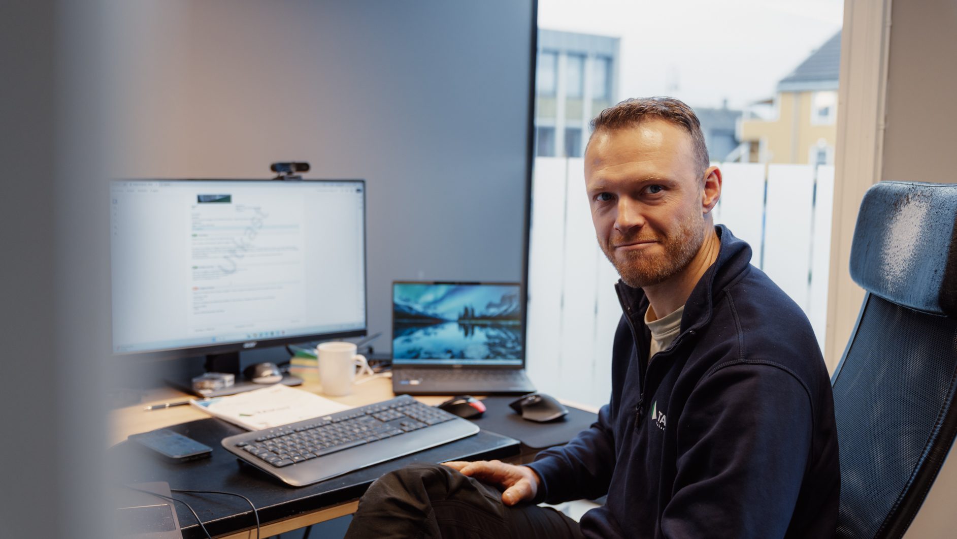Bearded man smiling at camera, sitting at an office desk with two monitors.