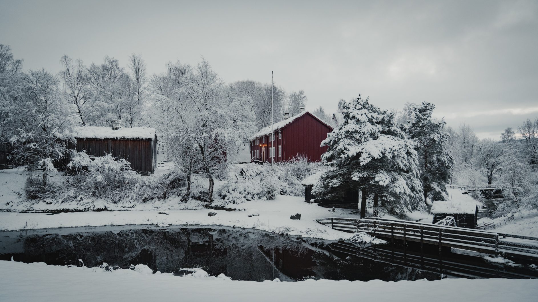A snowy winter scene with a red house, cabins, snow-covered trees, reflective water, and a small bridge.
