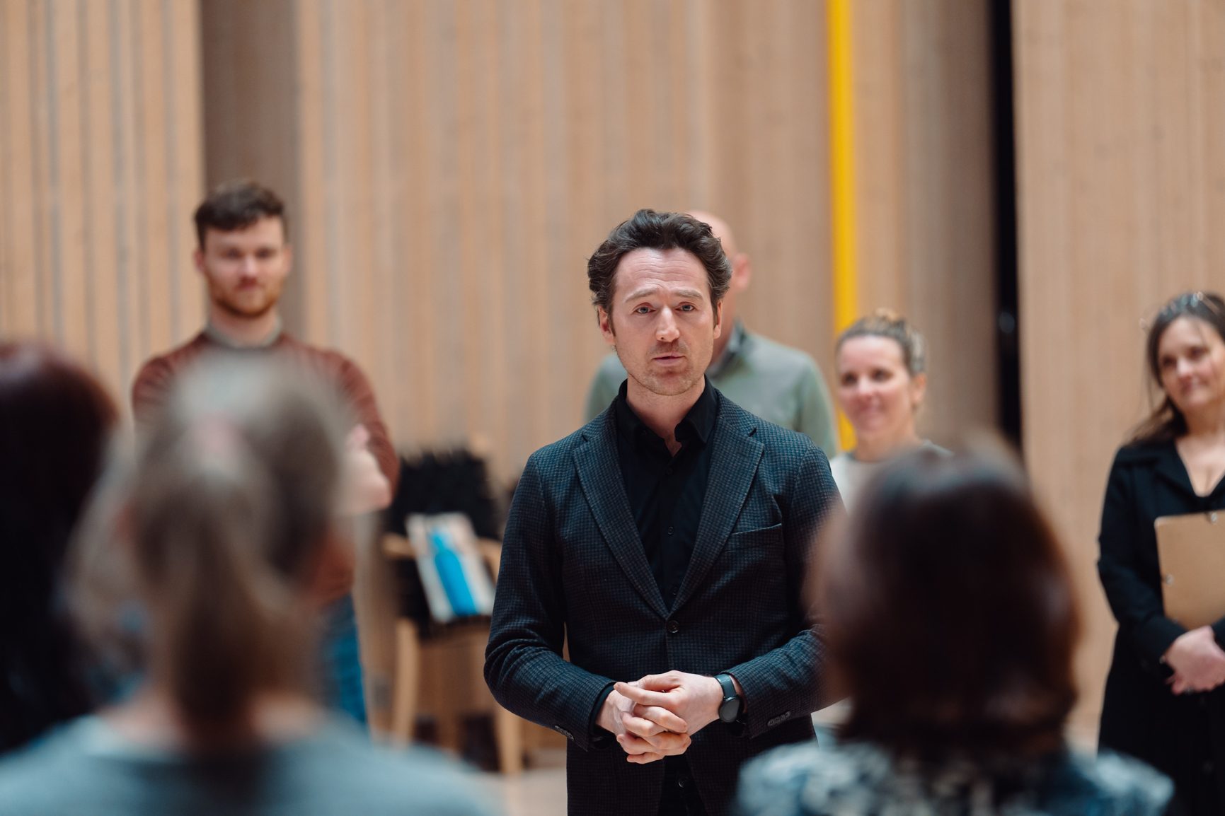 A man in a dark jacket addresses a diverse group of people in an indoor setting with wooden walls.
