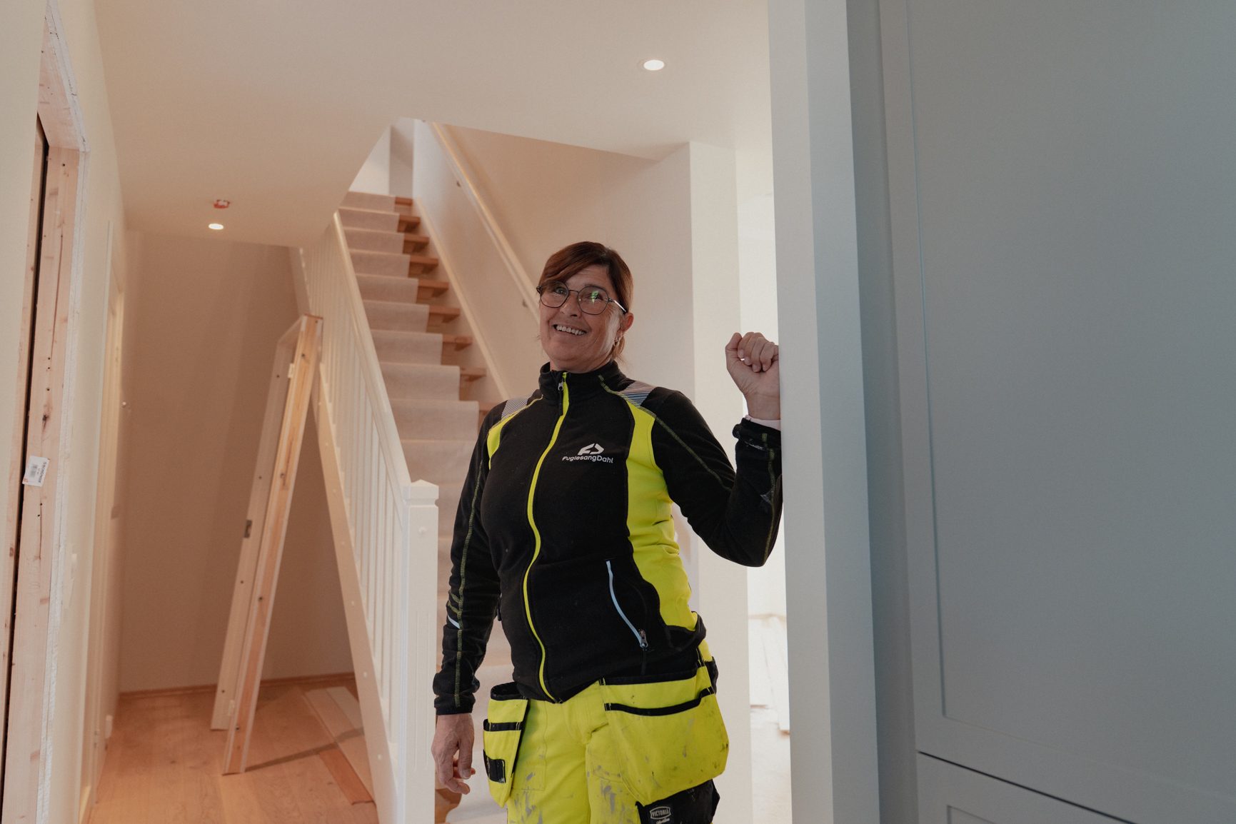 Smiling woman in work clothes leaning in a renovated house with a staircase in the background.