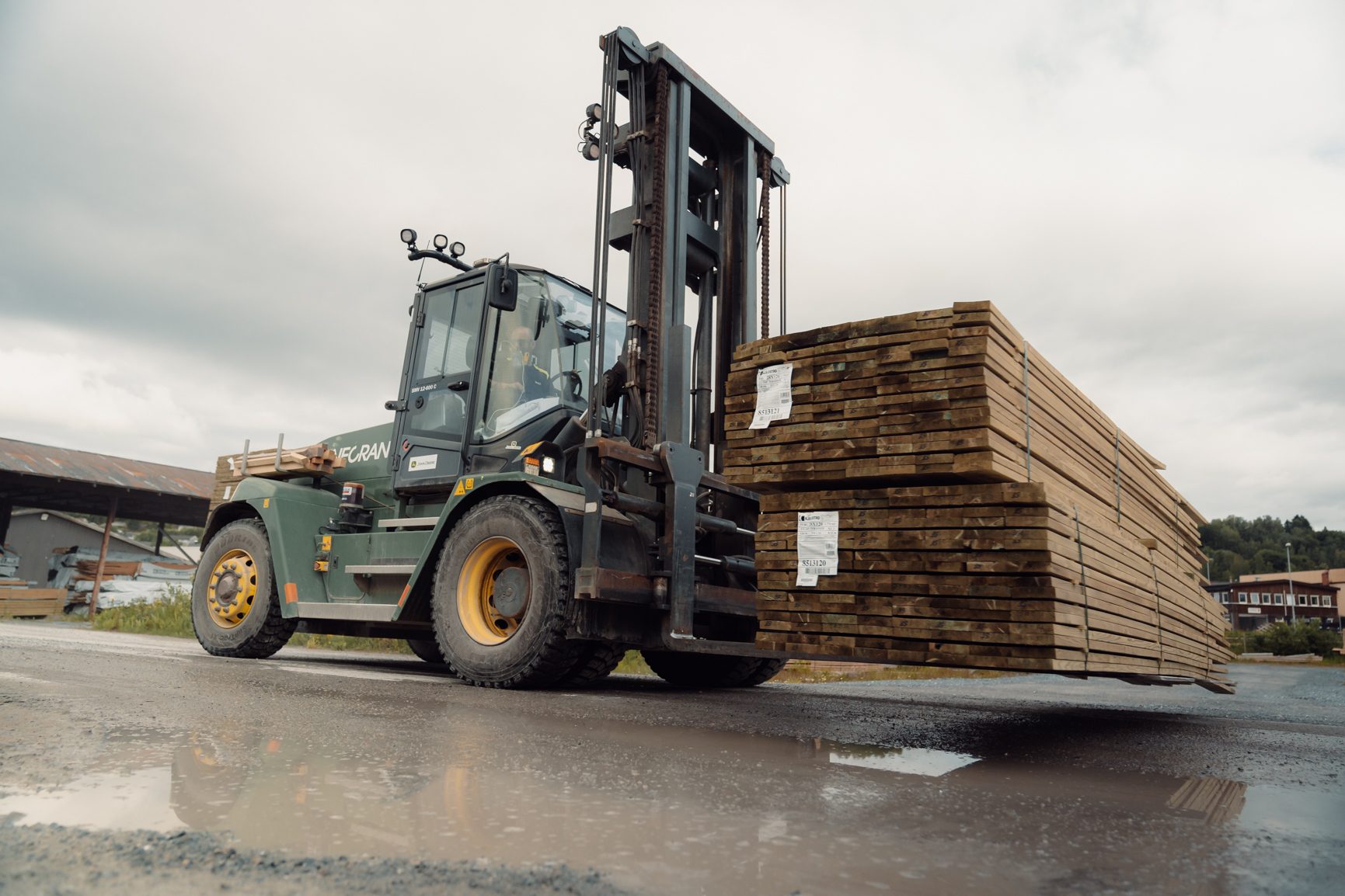 Heavy forklift moving large lumber stacks on a wet road.