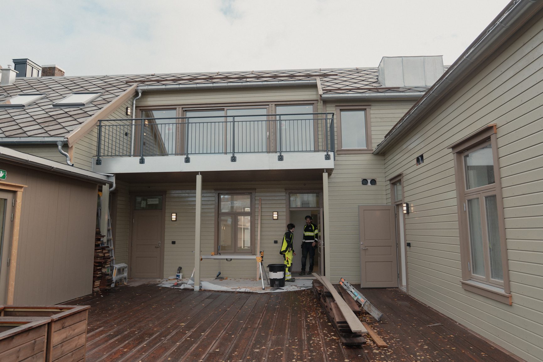 Exterior of a light-sided building with a wooden deck. Two workers are at an entrance below a balcony.
