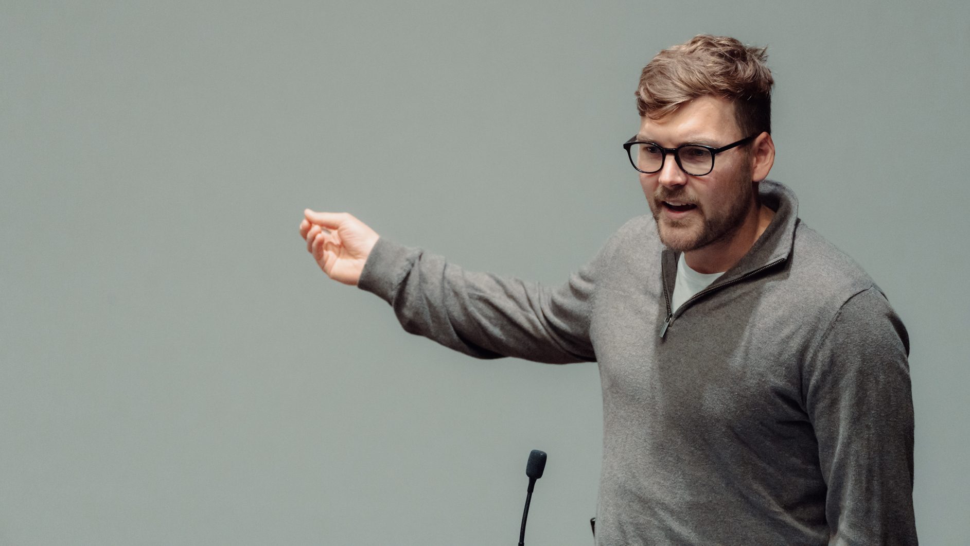 A man in glasses and a grey sweater speaks, gesturing, with a microphone visible below him.