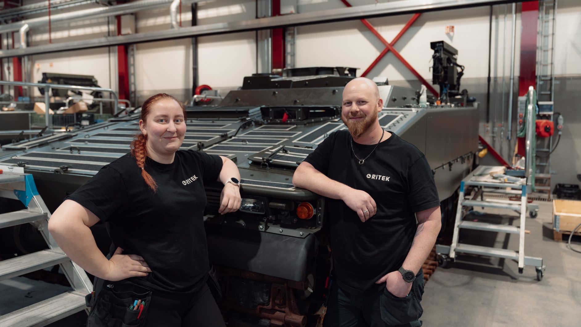 Two workers in QRITEK shirts pose with a military tank in a workshop.
