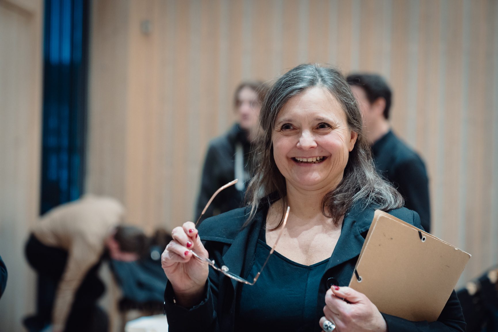 A smiling woman with graying hair holds glasses in one hand and a folder in the other, looking happily.