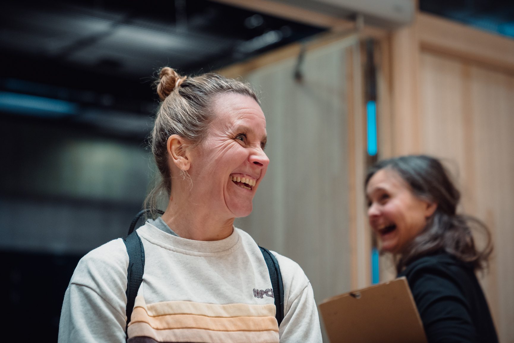 Two women laughing, one in the foreground with a hair bun and striped sweatshirt, another blurred in the background.