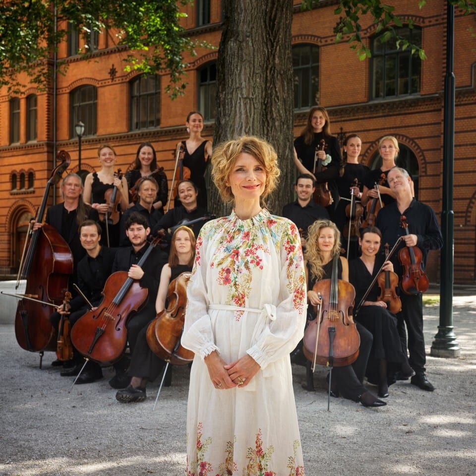 Smiling woman in floral dress, orchestra behind her outdoors.