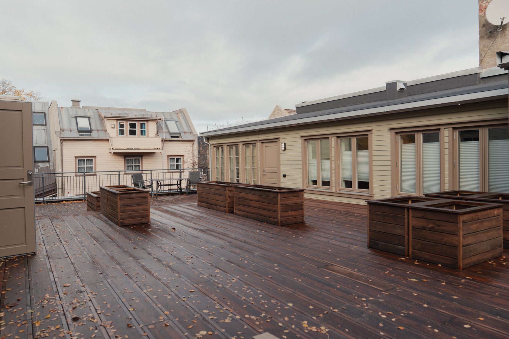 Wet wooden deck with empty planters and autumn leaves, surrounding buildings under an overcast sky.
