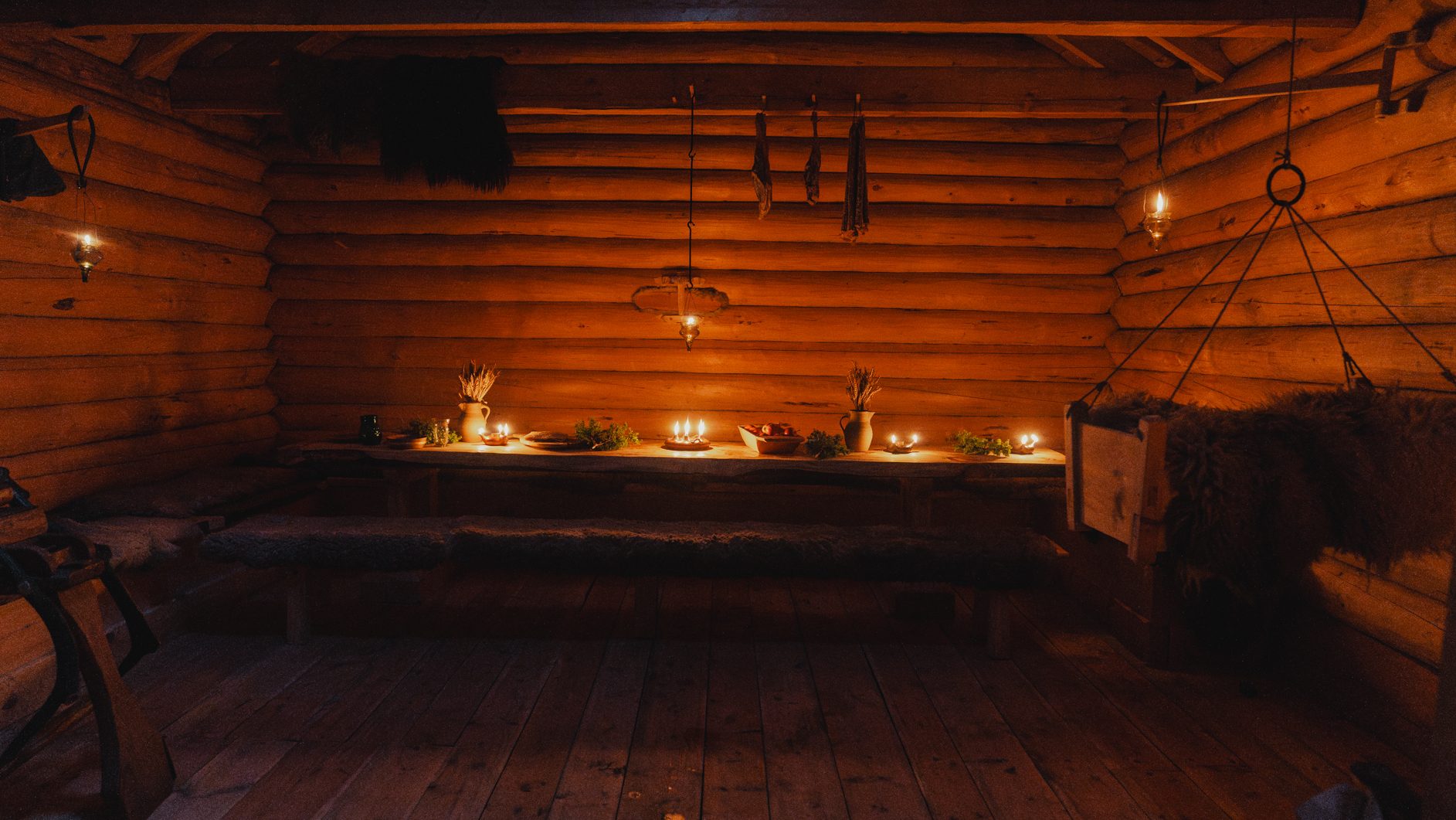 Cozy, candlelit log cabin interior with a long table, benches, and a hanging baby cradle.