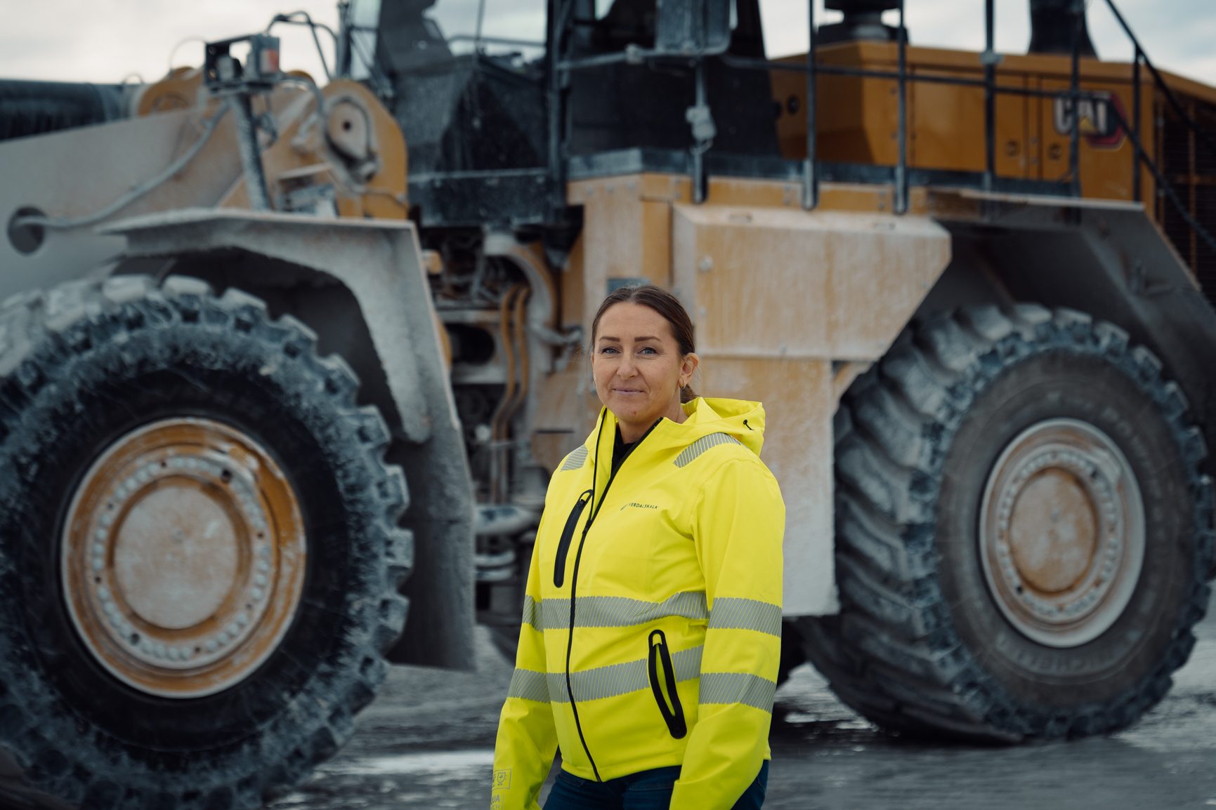 Woman in yellow safety jacket standing in front of large heavy construction equipment.