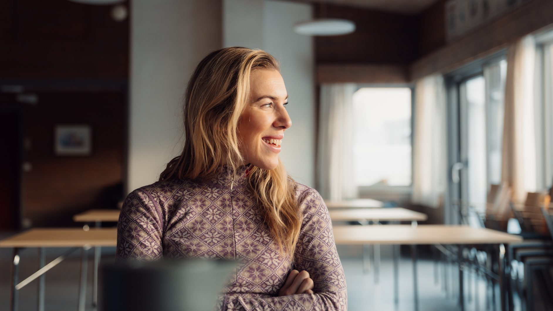 Happy blonde woman in a purple patterned top, looking right in a bright room.