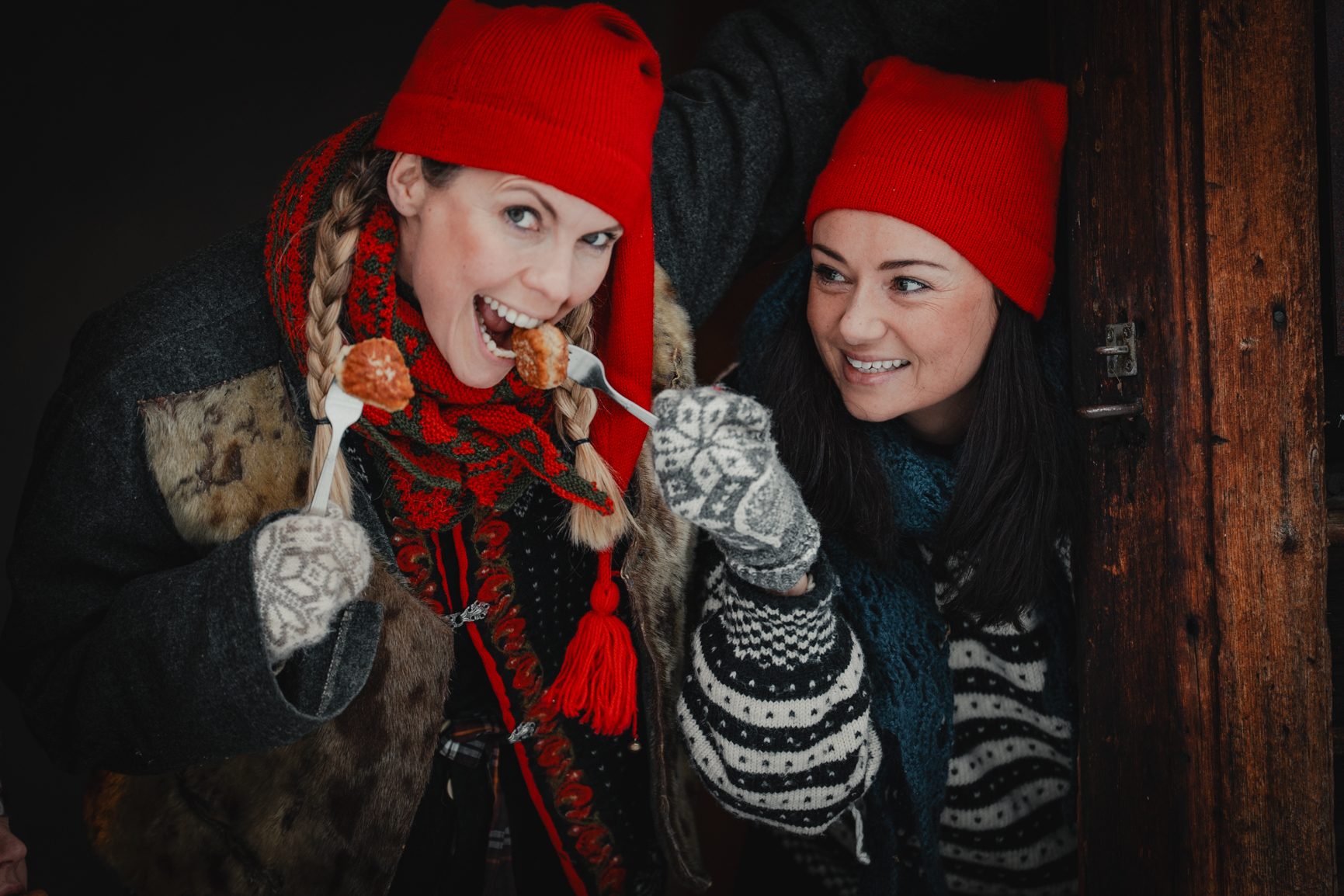 Two women in red hats and warm clothes. One eats from a fork, the other smiles.
