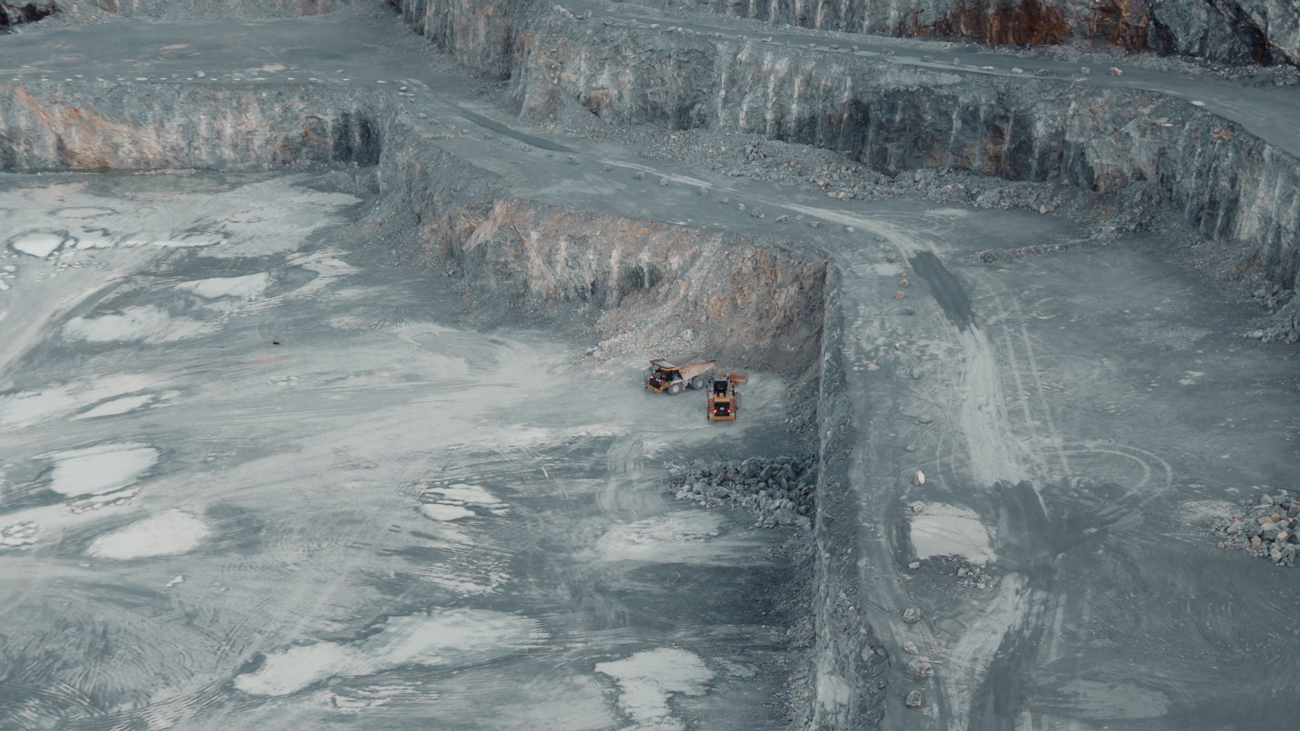 Aerial view of a large, grey quarry with rock walls and two mining vehicles.