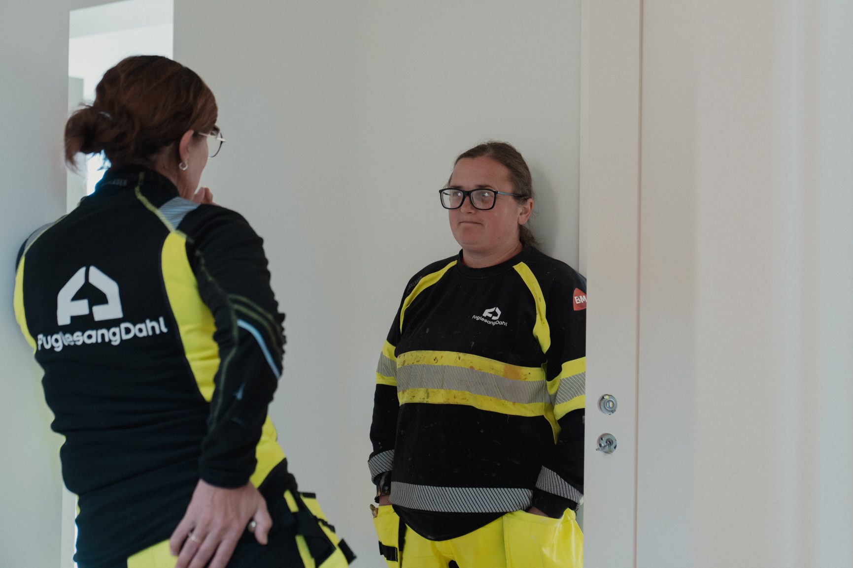 Two female construction workers in black and yellow uniforms with "PuglesangDahl" logos talk indoors.