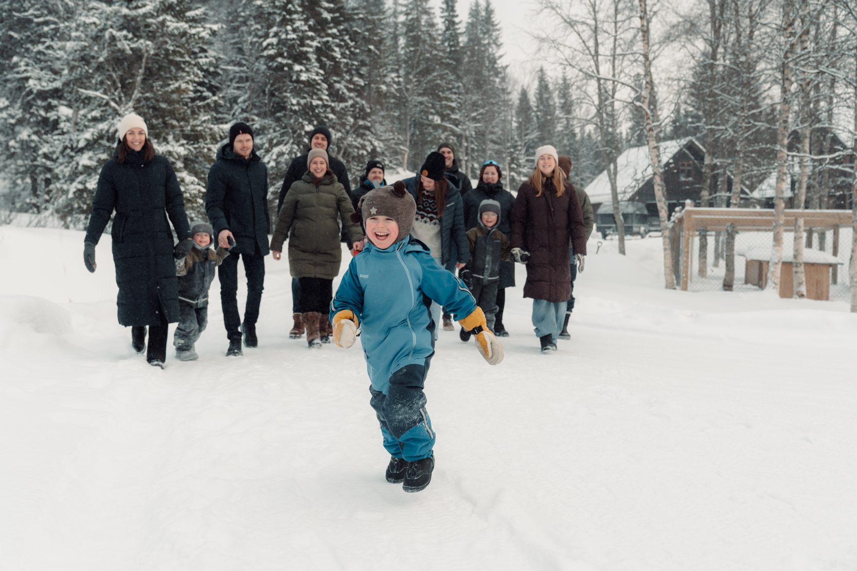 A happy child runs through deep snow, leading a group of adults and children in a winter forest.