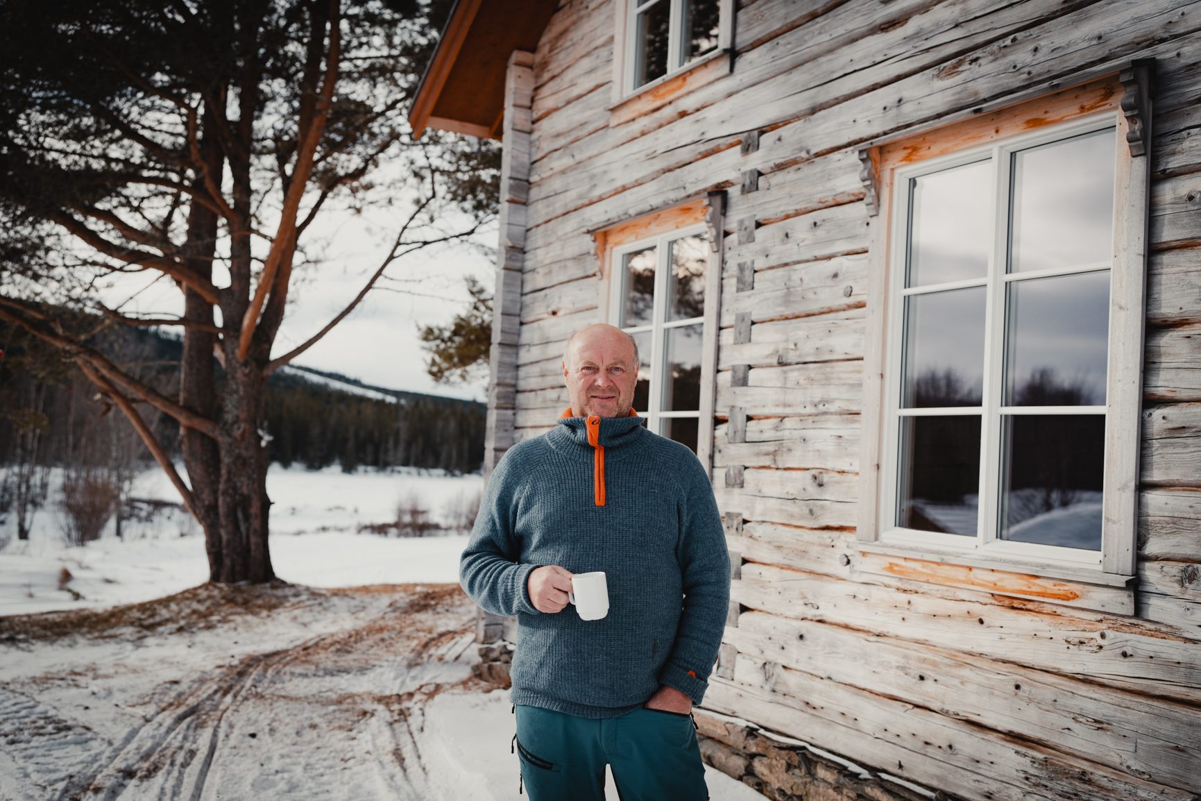Man in a blue sweater holding a mug by a rustic wooden cabin in a snowy winter landscape.