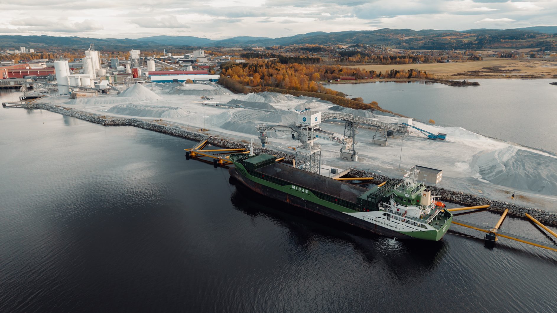 Aerial view of a cargo ship docked at an industrial port with material piles, silos, and an autumn landscape.