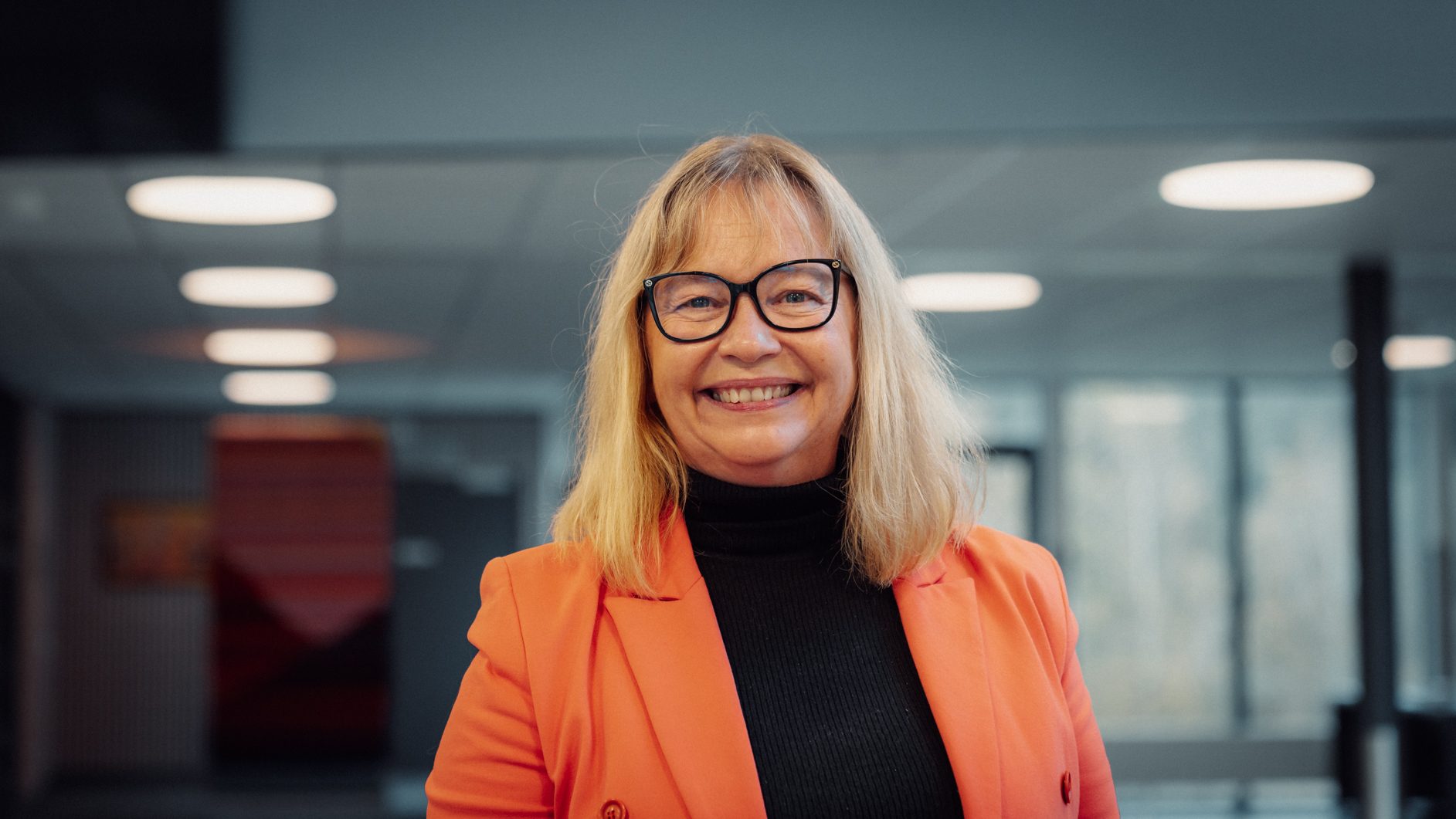Smiling blonde woman with glasses wearing an orange blazer.
